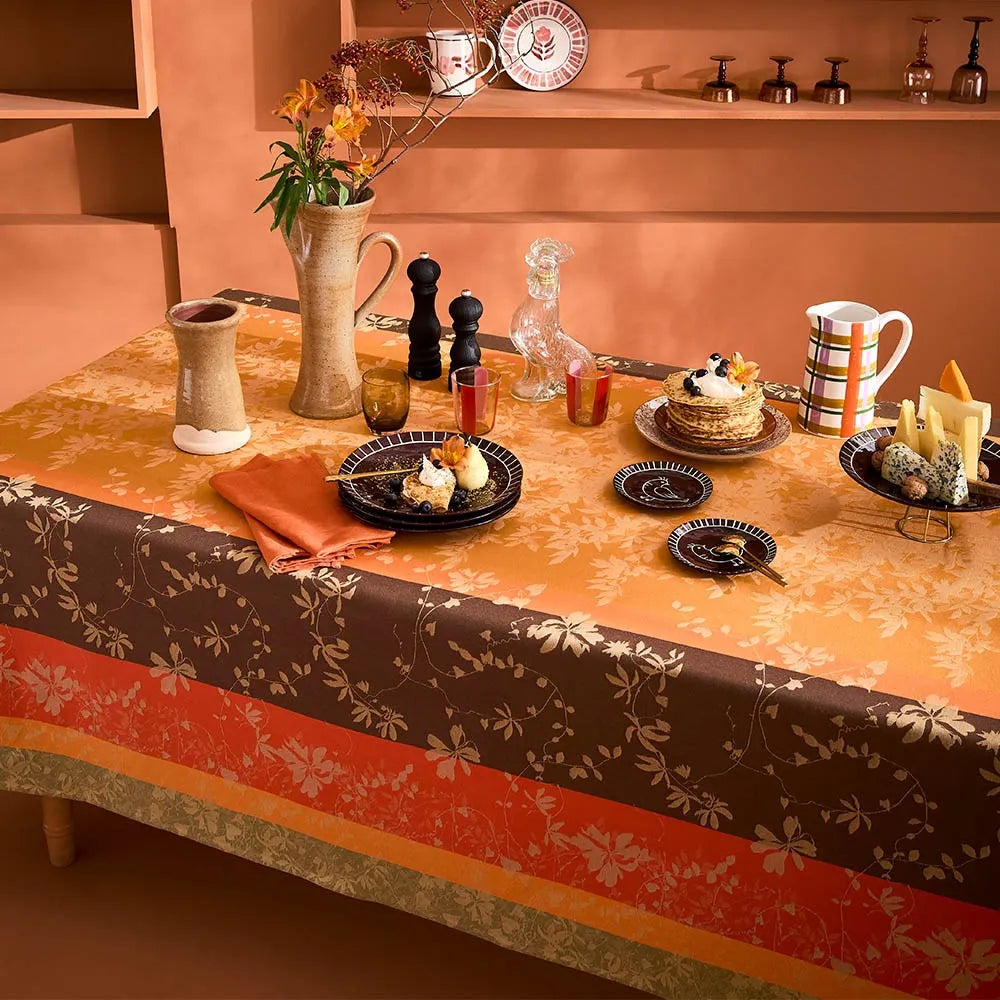A striped autumn leaf pattern tablecloth dressed on a set table, complete with dark wooden chairs and decorative plates displayed on wall shelving.
