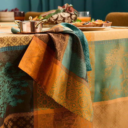 A woven cotton jacquard tablecloth and napkin featuring a warm patchwork plaid pattern of geometric squares in teal, orange, and amber.