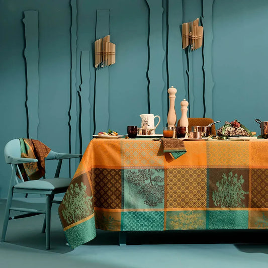 A woven cotton jacquard tablecloth featuring a warm, rich patchwork plaid pattern in shades of orange, teal, and amber, beautifully styled on a dining table against a textured teal wall.