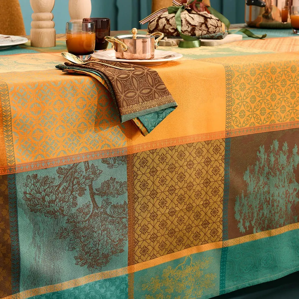 A woven cotton jacquard tablecloth featuring an autumnal patchwork layout of geometric squares and rectangular panels, filled with detailed evergreen tree motifs and intricate patterns in shades of orange, teal, and amber.