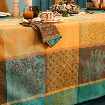 A woven cotton jacquard tablecloth featuring an autumnal patchwork layout of geometric squares and rectangular panels, filled with detailed evergreen tree motifs and intricate patterns in shades of orange, teal, and amber.