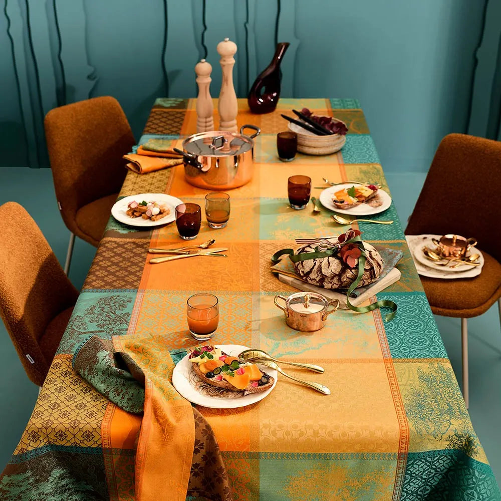 A woven cotton jacquard tablecloth featuring a warm patchwork plaid pattern of geometric squares in teal, orange, and amber, beautifully styled on a long dining table set with white dinner plates, elegant glassware, and a copper serving pot against a deep teal wall.