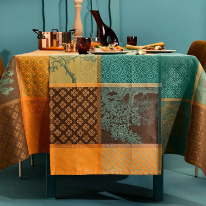A woven cotton jacquard table runner featuring a warm patchwork plaid pattern of geometric squares in teal, orange, and amber, beautifully styled on a table set with elegant glassware, and a copper serving pot against a deep teal wall.