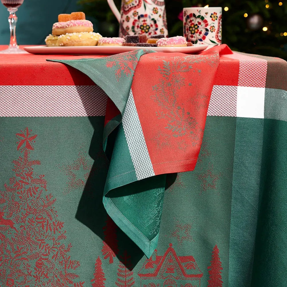 A festive Christmas-themed tablecloth in green and red, featuring a tree and house pattern, draped over a table with a matching folded napkin. In the background, a holiday tea set and pastries are visible on a blurred outdoor table surface.