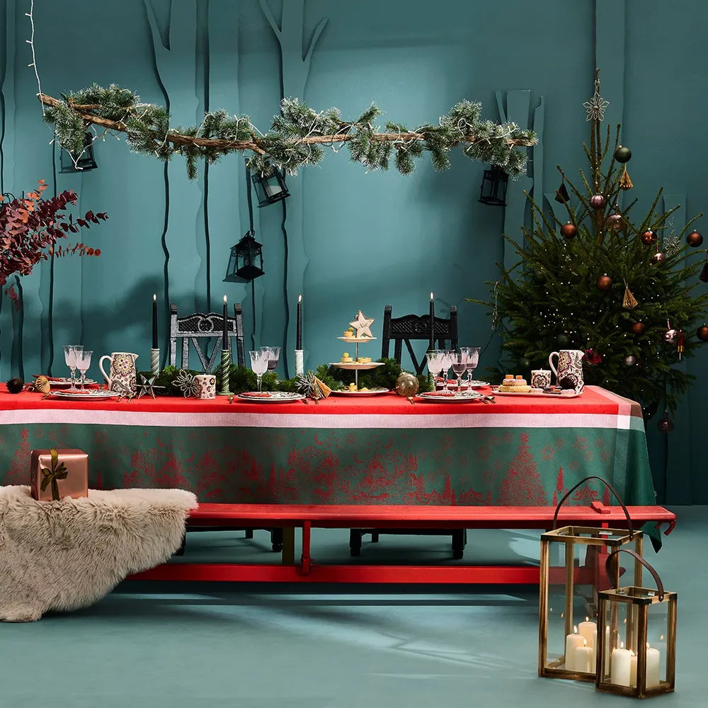 A beautifully set holiday table featuring the Noel Magique Houx green and red cotton jacquard tablecloth. The scene includes a red wooden bench draped with a cozy sheepskin rug, a decorated Christmas tree in the background, a festive garland hanging above with black lanterns, and elegant brass lanterns with glowing candles on the floor.