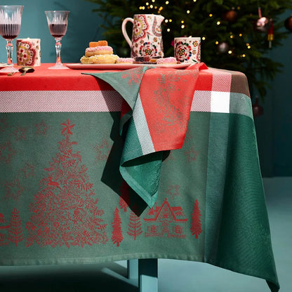 A holiday table setting featuring the Garnier-Thiebaut Noel Magique Houx green and red cotton jacquard tablecloth and matching napkin in a warm teal room with Christmas decorations. 