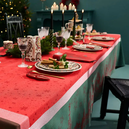 A festive holiday dining table styled with the Noel Magique Houx cotton jacquard tablecloth, showing its vibrant red top patterned with stars, a woven white-and-red border, and a green-and-red drop. The elegant setting features holiday plates with appetizers, crystal glasses, black taper candles, and miniature Christmas trees against a deep teal wall with a glowing fireplace mantel.