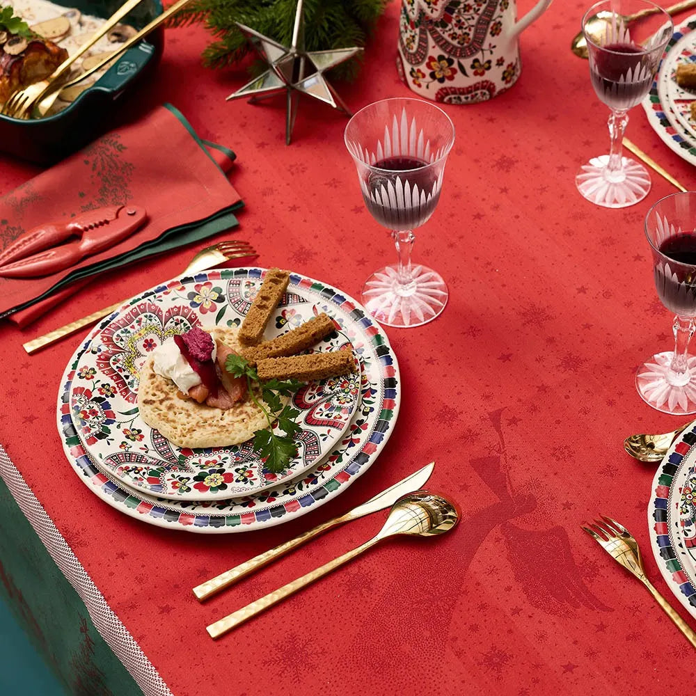 A close-up view of a holiday dining table set with the Noel Magique Houx red cotton jacquard tablecloth. The setting features decorative holiday plates with appetizers, polished gold flatware, crystal glasses filled with red wine, a matching red napkin, and a glimpse of a festive tea set on a beautifully patterned red table surface.
