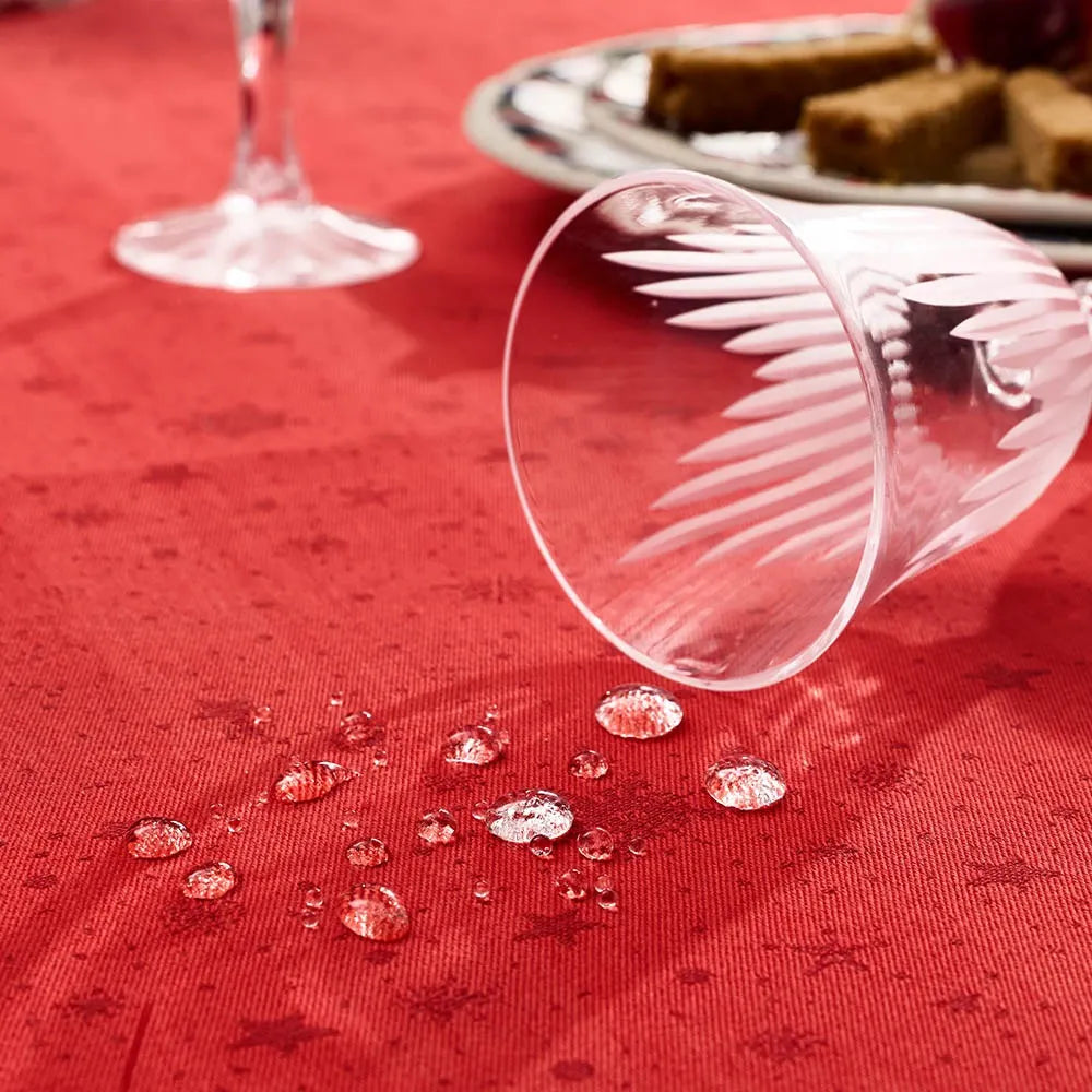 A close-up demonstrating the stain-resistant "Green Sweet" treatment on the Noel Magique Houx red cotton jacquard tablecloth. Liquid droplets bead up perfectly on the star-patterned fabric surface next to a fallen, empty crystal glass, with a plate of appetizers softly blurred in the background.