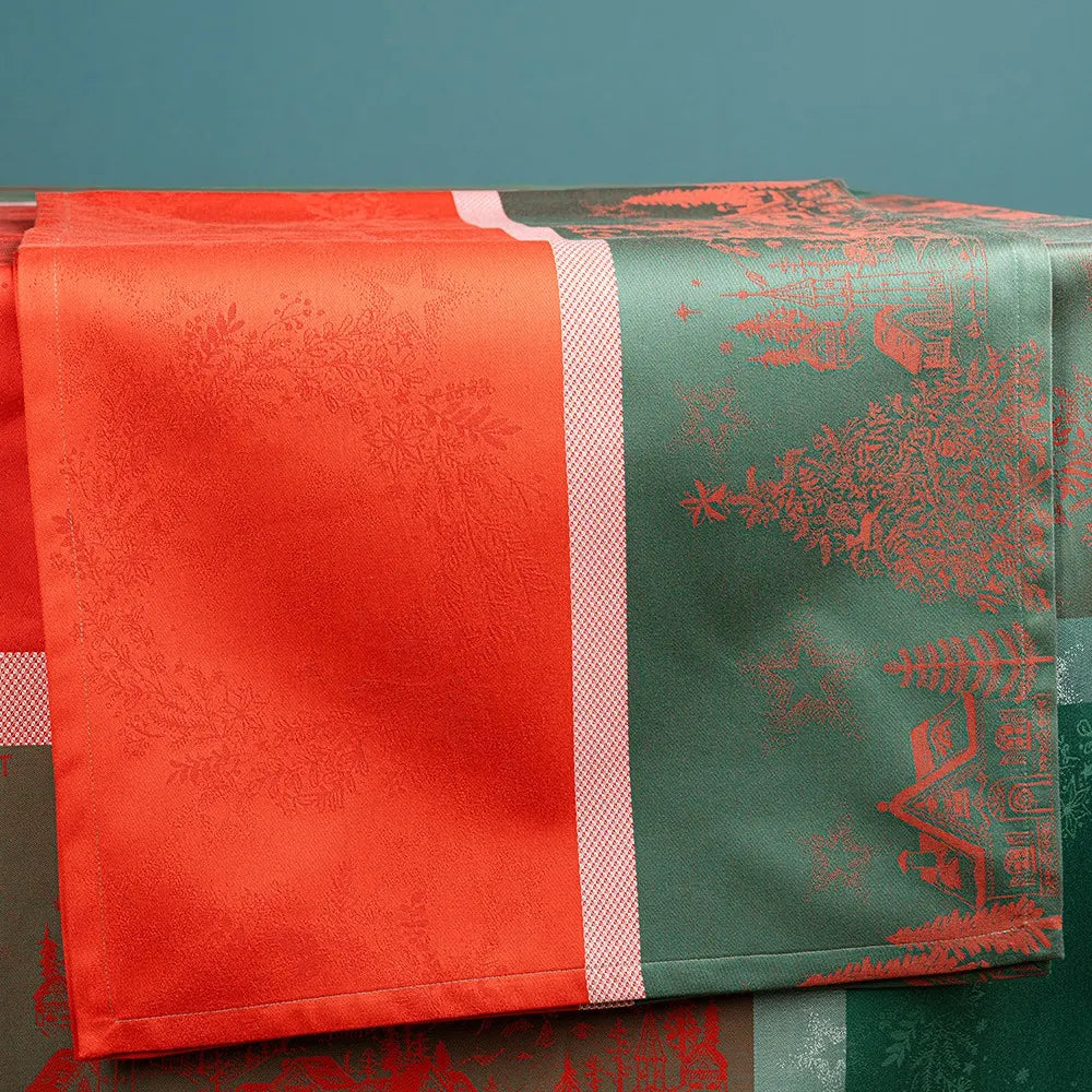 A close-up of the Noel Magique Houx cotton jacquard table runner draped over a surface, highlighting its split-panel festive design. The left panel is a vibrant red woven with subtle holly wreath patterns, while the right panel is a forest green featuring detailed red illustrations of Christmas trees and houses, separated by a textured pink and white stripe.