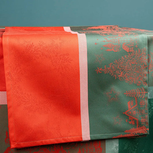 A close-up of the Noel Magique Houx cotton jacquard table runner draped over a surface, highlighting its split-panel festive design. The left panel is a vibrant red woven with subtle holly wreath patterns, while the right panel is a forest green featuring detailed red illustrations of Christmas trees and houses, separated by a textured pink and white stripe.