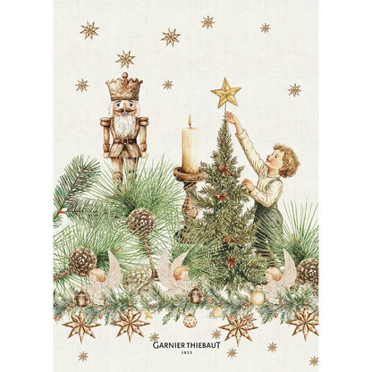 A Garnier-Thiebaut cotton-linen kitchen towel with a vintage illustration, holiday design. The print features a classic wooden nutcracker, a child placing a golden star on a Christmas tree, lit candles, pine cones, and scattered gold stars.