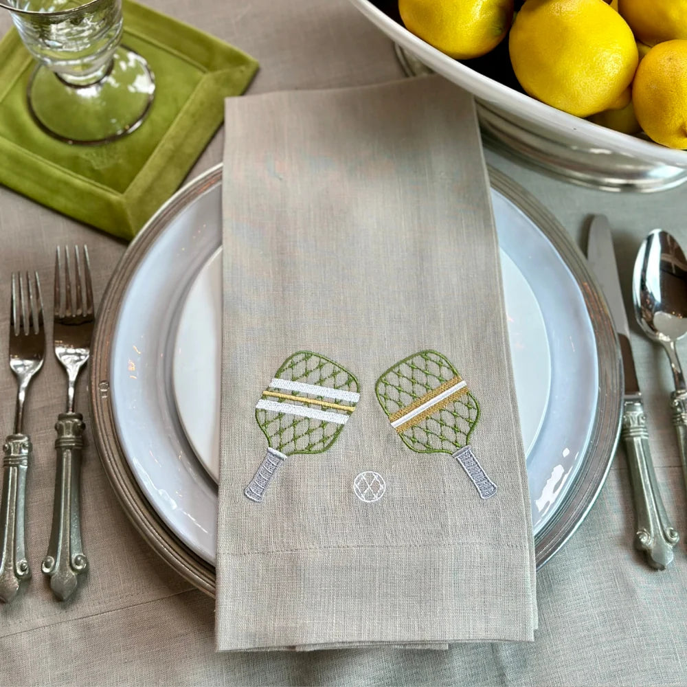 Flax colored linen Napkin with pickleball paddles embroidery on a table setting with plates, cutlery, and lemons.