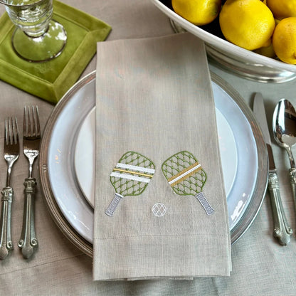 Flax colored linen Napkin with pickleball paddles embroidery on a table setting with plates, cutlery, and lemons.