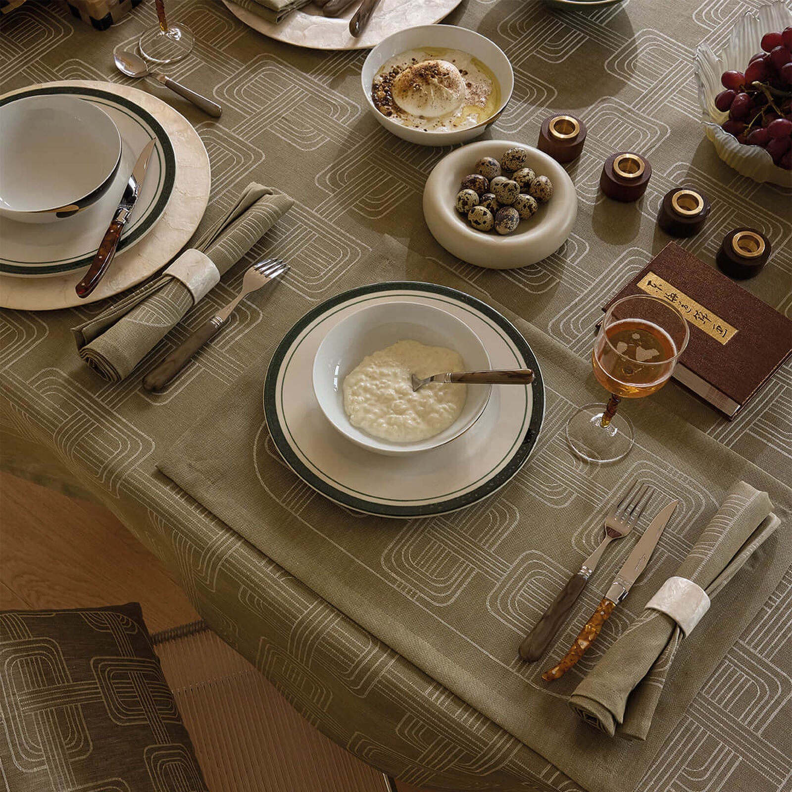 High-angle view of a dining table set with an olive green Jacquard linen tablecloth and matching napkins, styled with grey-rimmed white bowls, bowls of yogurt and quail eggs, and a glass of rosé.