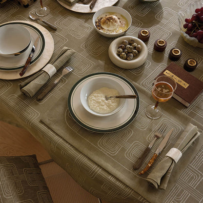 High-angle view of a dining table set with an olive green Jacquard linen tablecloth and matching napkins, styled with grey-rimmed white bowls, bowls of yogurt and quail eggs, and a glass of rosé.