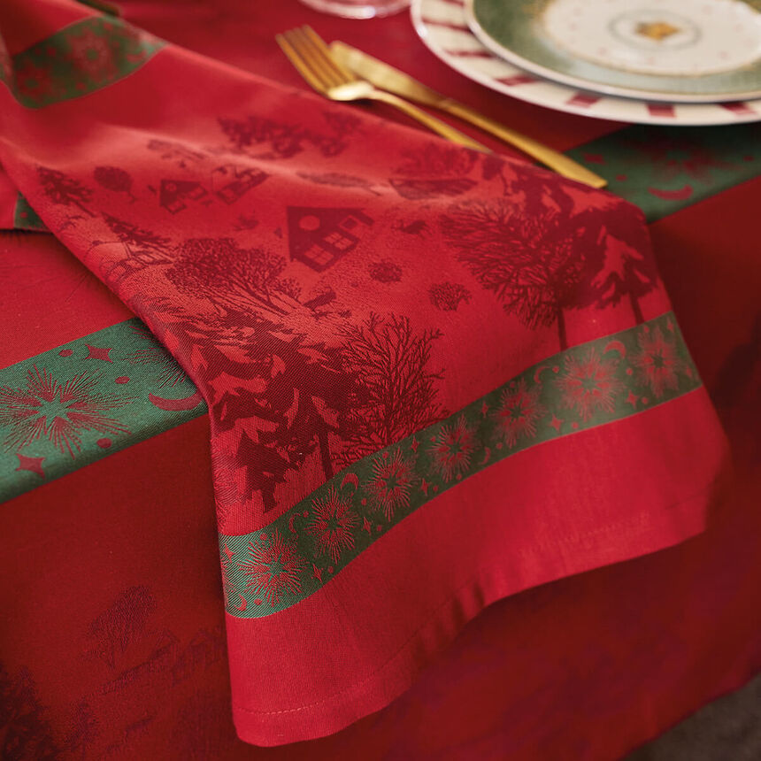 Detail view of a red cotton jacquard tablecloth and matching napkin featuring a winter village and tree pattern, accented by a deep green patterned border stripe with gold cutlery on top.