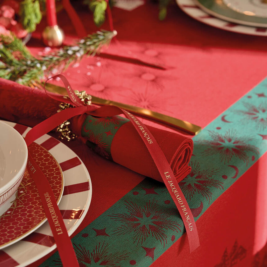 A festive table setting with a red tablecloth, a rolled red napkin tied with a red ribbon printed with 'LE JACQUARD FRANCAIS', gold-toned flatware, patterned plates, and a teal runner with red floral motifs.