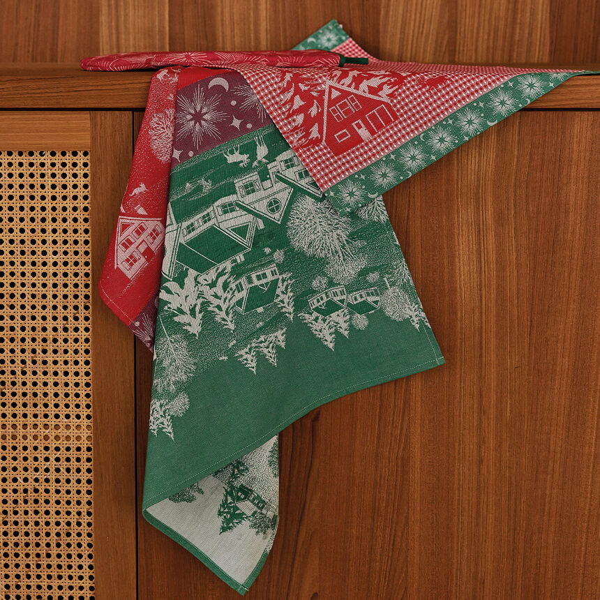 A dishtowel with a Christmas village pattern, draped over a wooden cabinet. The fabric features a green section with white winter house illustrations, a red gingham section with white snowflakes, and a red section with white floral motifs.