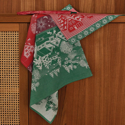 A dishtowel with a Christmas village pattern, draped over a wooden cabinet. The fabric features a green section with white winter house illustrations, a red gingham section with white snowflakes, and a red section with white floral motifs.