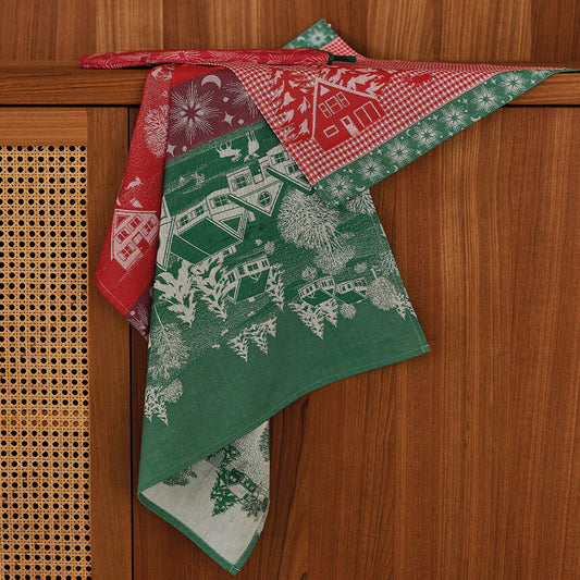 A dishtowel with a Christmas village pattern, draped over a wooden cabinet. The fabric features a green section with white winter house illustrations, a red gingham section with white snowflakes, and a red section with white floral motifs.