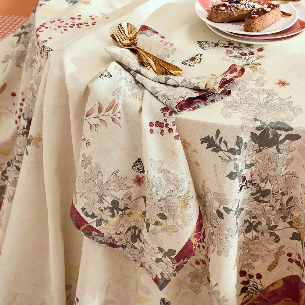 A cream-colored tablecloth with a detailed botanical and butterfly pattern, styled on a dining table with a matching folded napkin, gold cutlery, and a plate of food.