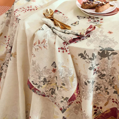 A cream-colored tablecloth with a detailed botanical and butterfly pattern, styled on a dining table with a matching folded napkin, gold cutlery, and a plate of food.