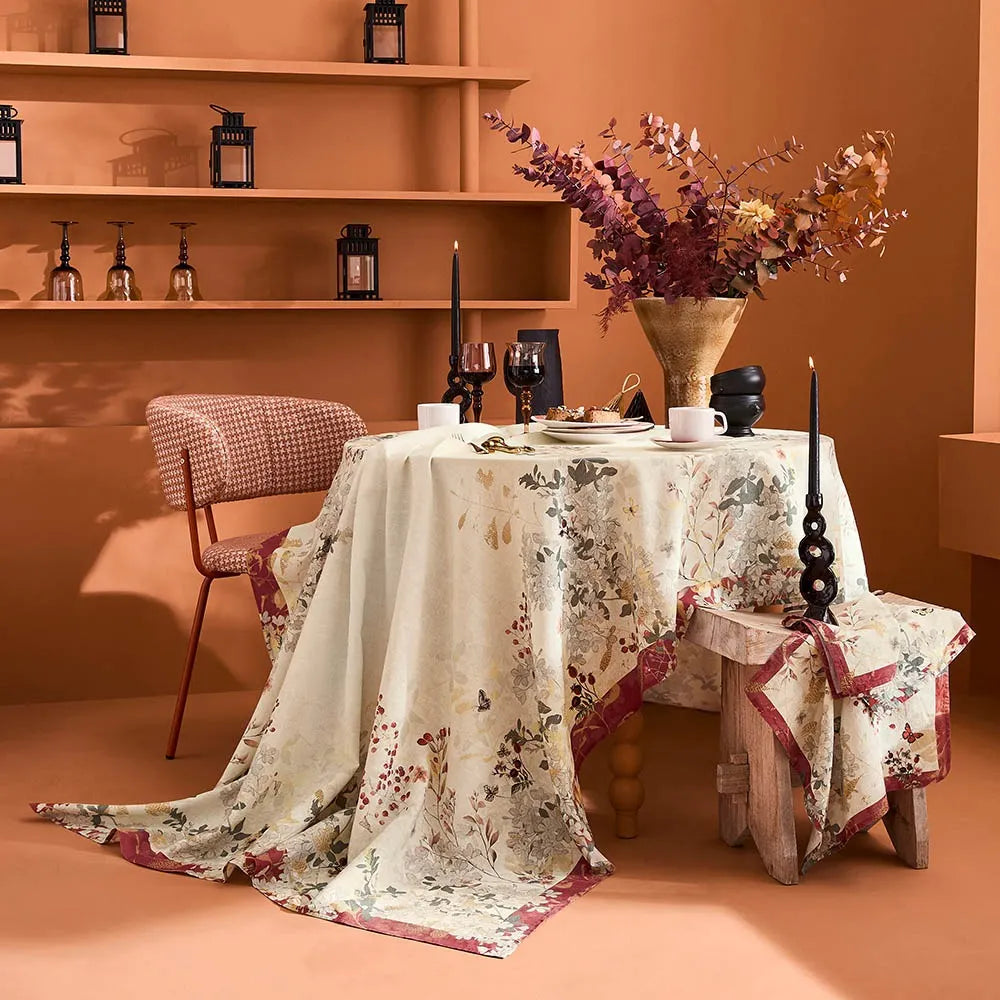 A styled dining table in a warm terracotta room, covered with a cream cotton-linen tablecloth featuring a detailed vintage floral print with red berries and muted green foliage, beautifully set with modern white cups, elegant wine glasses, and a large golden vase of dried plum-colored florals.