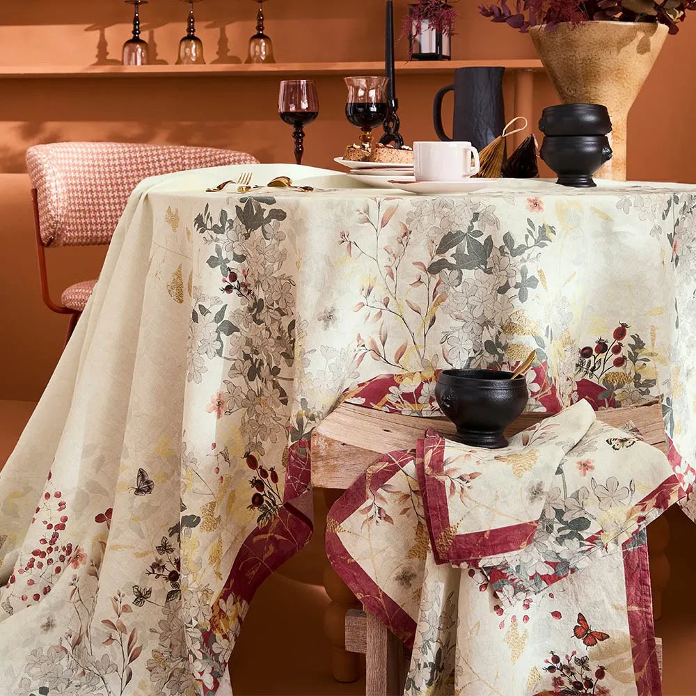 A cranberry and floral tablecloth draped over a dining table in a warm, rustic room setting, featuring a cream base with red, green, and gray botanical patterns.