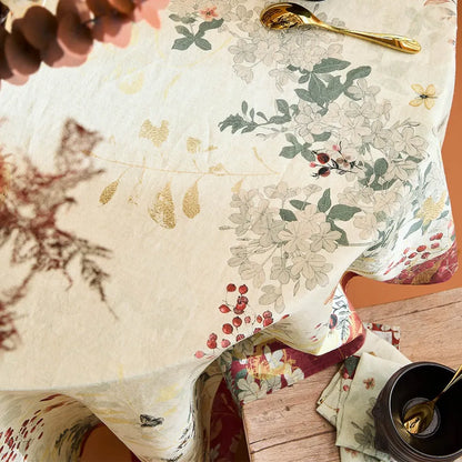 A close-up of a floral tablecloth with a cream base, green leaves, and muted red berry accents, styled on a wooden table with a gold spoon and a dark cup.