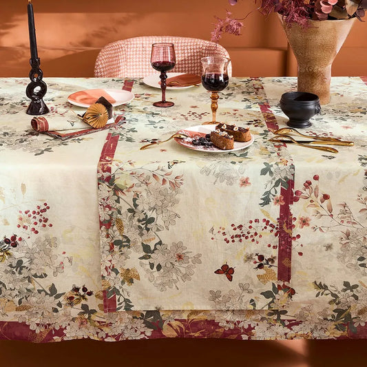 A floral tablecloth and matching table runner with a cream base and red, green, and beige botanical print, styled on a dining table with wine glasses, plates, candles, and a vase in a warm, rustic setting.