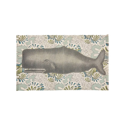 A top-down view of a rectangular, hand-loomed cream cotton bath rug featuring a large-scale illustration of a majestic charcoal gray whale set against an intricate, vintage botanical floral pattern background in soft sage green and denim blue tones.