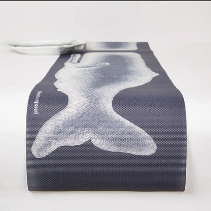 A detailed, close-up view of a gray woven vinyl table runner featuring a large, hand-screened illustration of a majestic whale tail on a classic basketweave texture, styled with a white ceramic plate on a table.
